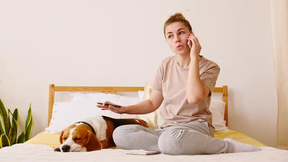 Young Woman Sits on the Bed and Petting Her Dog Beagle While Talking By Phone