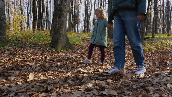 In slow motion, mother and daughter holding hands walk and kick fallen leaves in an autumn forest