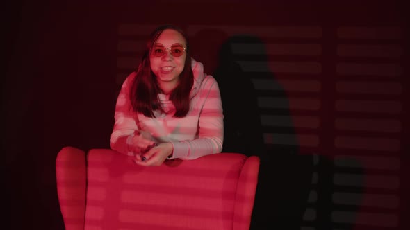Young Woman in Glasses Standing Behind Armchair in Dark Room with Falling Red Light and Shadow