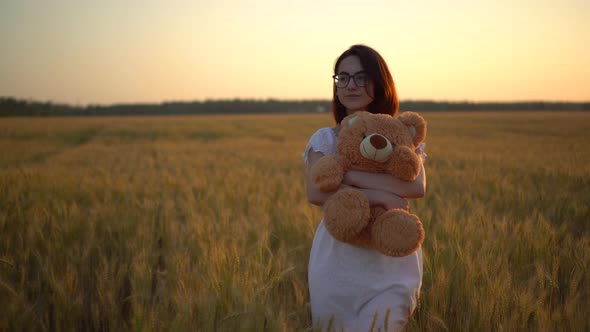 A Young Woman Walks Through a Wheat Field with a Teddy Bear at Sunset. Girl Hugs a Teddy Bear in