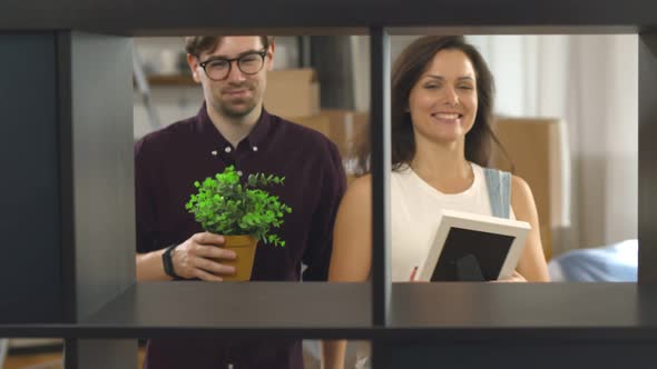 Young Couple in Love Putting Pot with Plant and Photo Frame on Shelves and Hugging