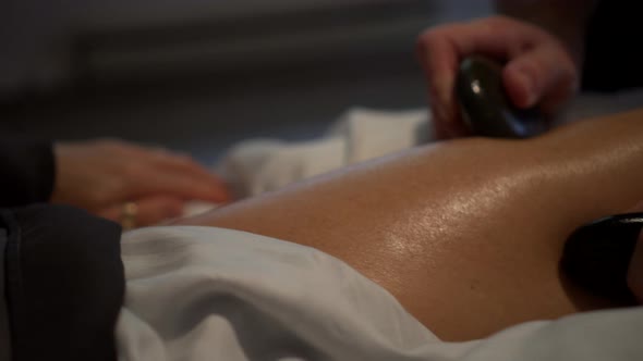 Very tight shot of massage therapist using two smooth black stones in a circular motion on the thigh