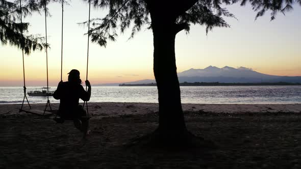 Beautiful young woman swinging on a swing at majestic sunset tropical beach.