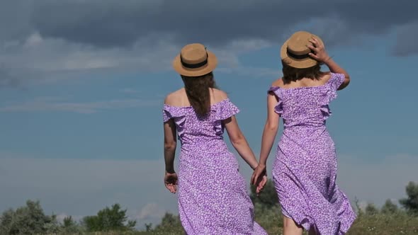Young Twins Sisters in Purple Summer Dresses Walks Green Field Holding Hands