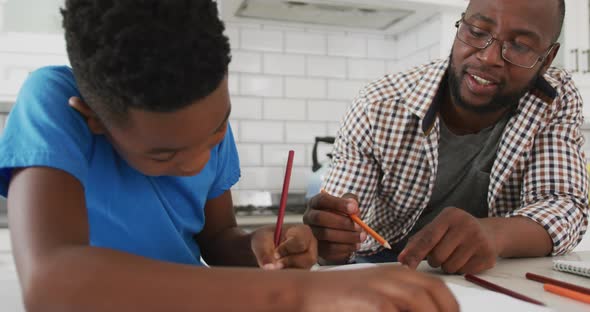 Happy african american father sitting at kitchen table helping son with school work