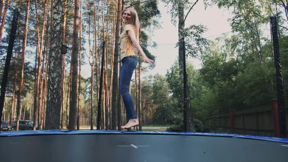 Happy Female Jumping on Trampoline. Beautiful Young Blond Barefoot Woman in Light Summer Shirt and