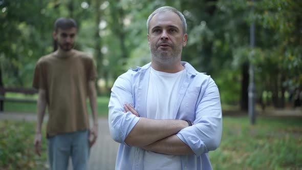 Portrait of Stubborn Middle Aged Father Standing in Park with Crossed Hands As Blurred Nervous Son