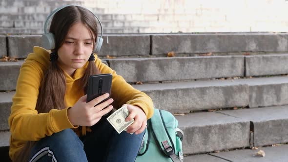 The Teenager Holds Dollar Bills and Looks at the Smartphone Screen