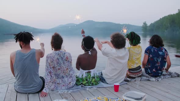 Friends Sitting on Pier and Waving Sparklers at Lake Party