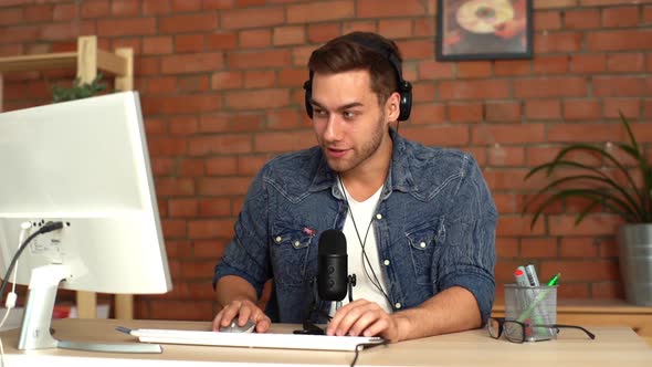 Cheerful Handsome Male Audio Blogger Wearing Headphones with Computer and Professional Microphone