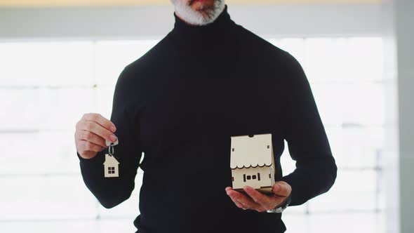 Male Real Estate Agent Raises His Hands with a Wooden House and Keys with a Keychain House Closeup