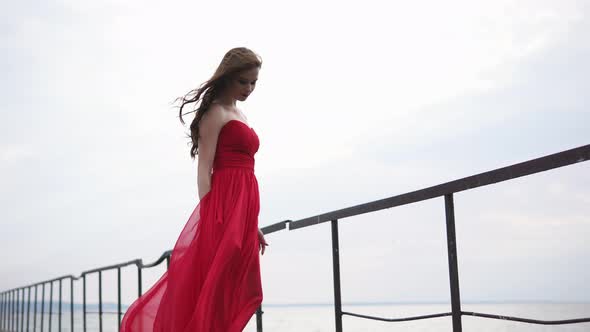 Beautiful Woman in Long Red Dress is Standing on a Sea Pier in Windy Weather