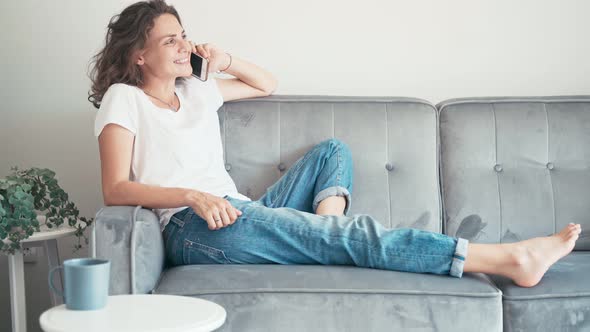 Young Cheerful Woman Talking on the Phone While Sitting on a Grey Couch