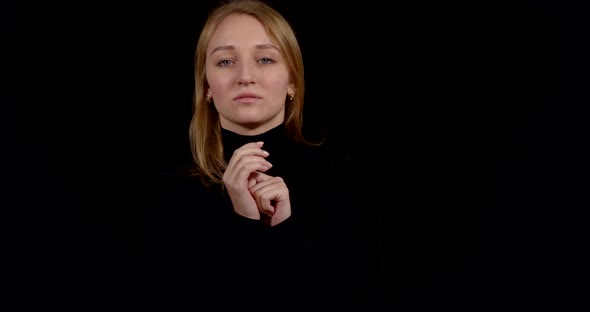 Charming Blonde Woman Is Standing at Dark Background, Wearing Black Gown, Folding Hands Near Chest