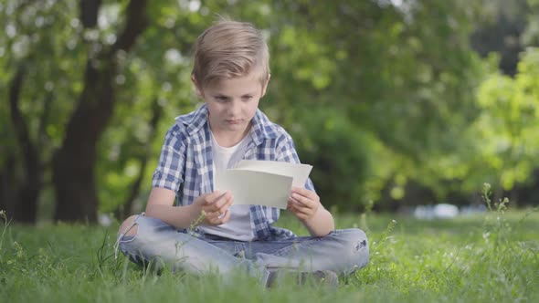 Portrait of Adorable Cute Boy in Checkered Shirt Looking at the Sheets of Paper in the Park