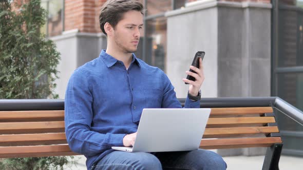 Young Man Using Smartphone and Laptop, Sitting on Bench