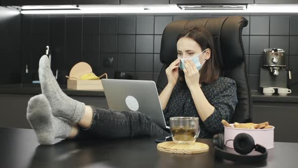 Young woman puts on protective medical face mask and starts working on laptop computer at home.