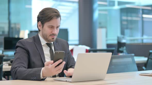 Businessman Browsing Internet on Smartphone While Using Laptop in Office