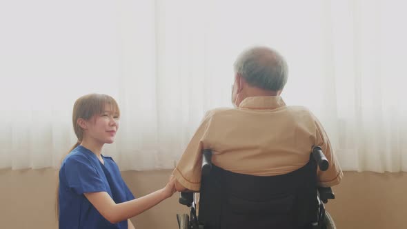 Asian nurse taking care of mature senior male patient sitting on wheelchair in hospital ward.