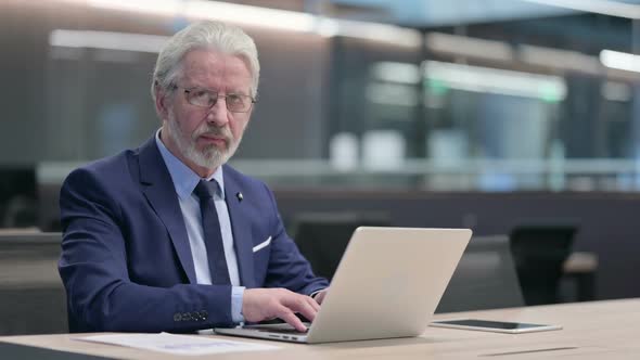 Old Businessman with Laptop Pointing at Camera Inviting