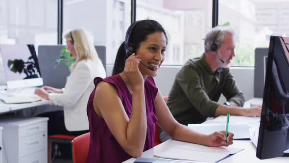 Mixed race businesswoman sitting using computer talking on phone headset in office