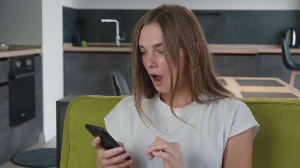 Worried Woman Reading Bad News in Message on Her Smartphone While Sitting on Sofa at Home. Shocked