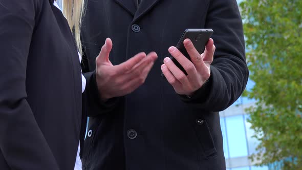 A Businessman and a Businesswoman Work on a Smartphone in Front of an Office Building - Closeup