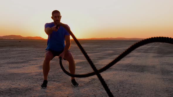 Athletic man working out with battle ropes on a dry lake at sunset