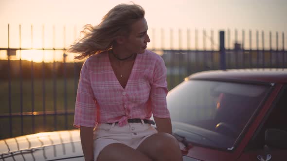 Beautiful Blonde Young Woman Sits on the Hood of Her Car at Sunset