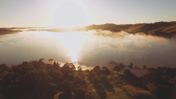 Aerial over a forest and lake covered in fog during the first morning light