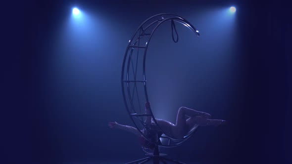 Gymnast Gracefully Moves the Body Lying on a Rotating Structure. Blue Smoke Background