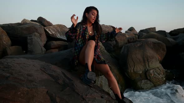 a Brunette in a Swimsuit Shirt and Rough Shoes is Sitting on Large Rocks on the Seashore