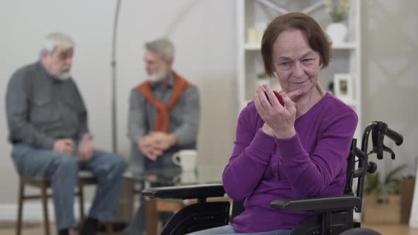 Positive Elderly Caucasian Woman Looking at Hand Mirror and Smiling As Men Chatting