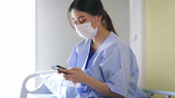 Young Asian Female Patient Sitting on Hospital Bed Wearing Surgical Face Mask Using Smartphone