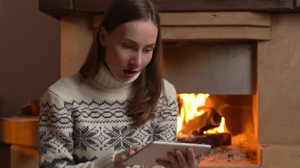 An Excited Woman Holds a Tablet and Celebrates the News While Sitting By the Fireplace