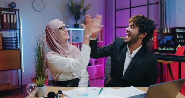 Male and Female Business Persons Sitting in front of Camera in Evening Workroom and Giving High Five