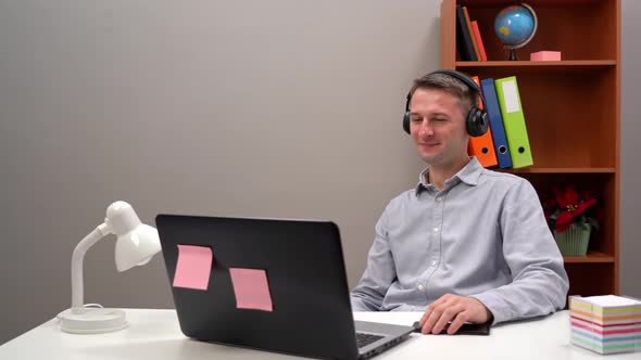 A Young Worker Communicates with a Client at an Online Conference From the Office Through Headphones