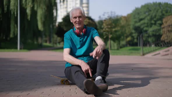 Wide Shot Confident Happy Senior Man Sitting on Skateboard Gesturing Horns with Fingers Smiling