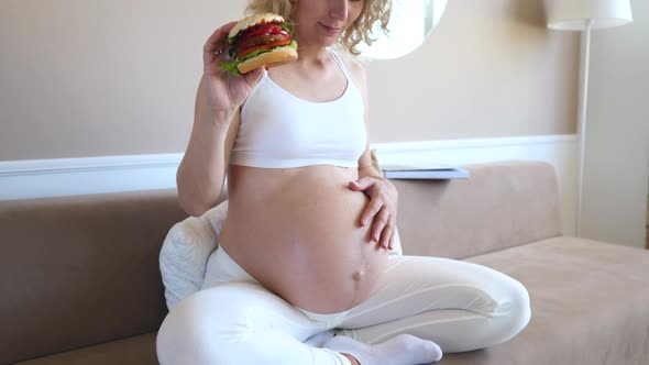 Hungry Pregnant Girl With Hamburger Sitting On Couch At Home