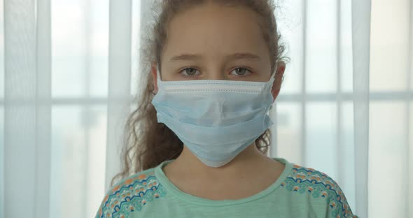 A Child at Home in a Protective Medical Mask with a Sad Face and Eyes Looks at the Camera