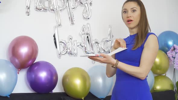 Surprised Young Woman Opening a Birthday Gift at Birthday Party with Colorful Balloons
