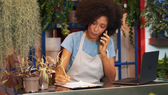 Smiling African female florist talking by phone and writing