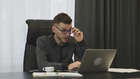 Exhausted young businessman working on laptop in office wearing glasses. Corporate business concept