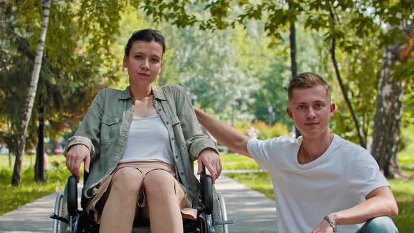 A Woman in a Wheelchair and a Man Sitting Next to Her in the Park  Looking in the Camera