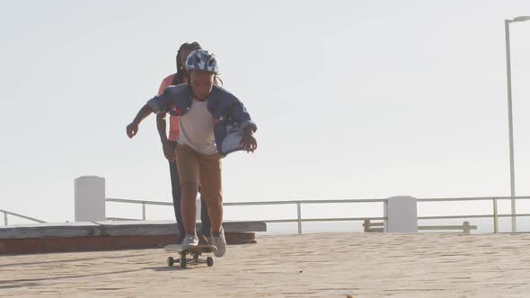 Video of happy african american father learning son how to skateboard on promenade