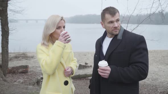 Cute Caucasian Couple Drinking Coffee and Talking on Riverbank