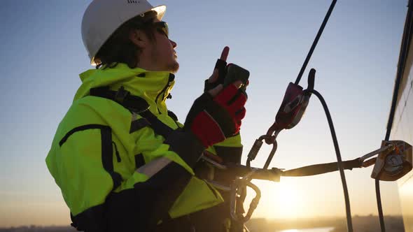 Man a Builder Engineer in a Helmet Hangs on Ropes on a High House with a Telephone and Conducts a