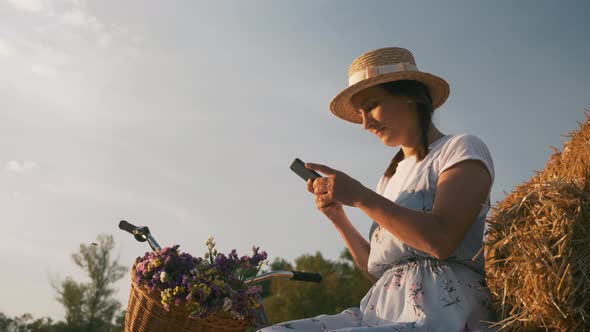 Young woman with mobile phone in hands is sitting on straw at sunset.