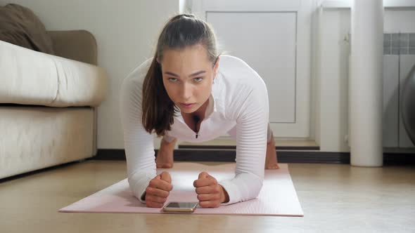 Woman Stands in Plank Pose Watching Tutorial on Mat in Room