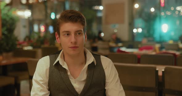 Serious Young Man is Sitting in a Restaurant Waiting Looking Around